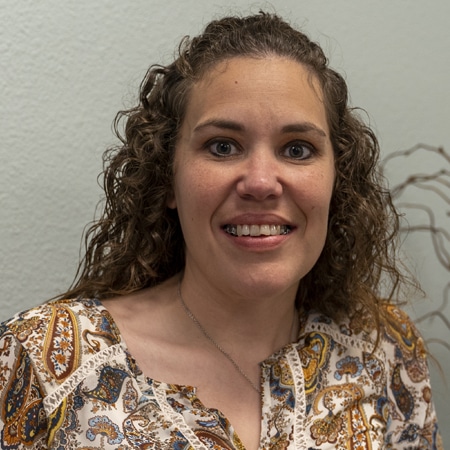 A woman with curly hair smiling in front of a plant, possibly affected by TMJ disorder.