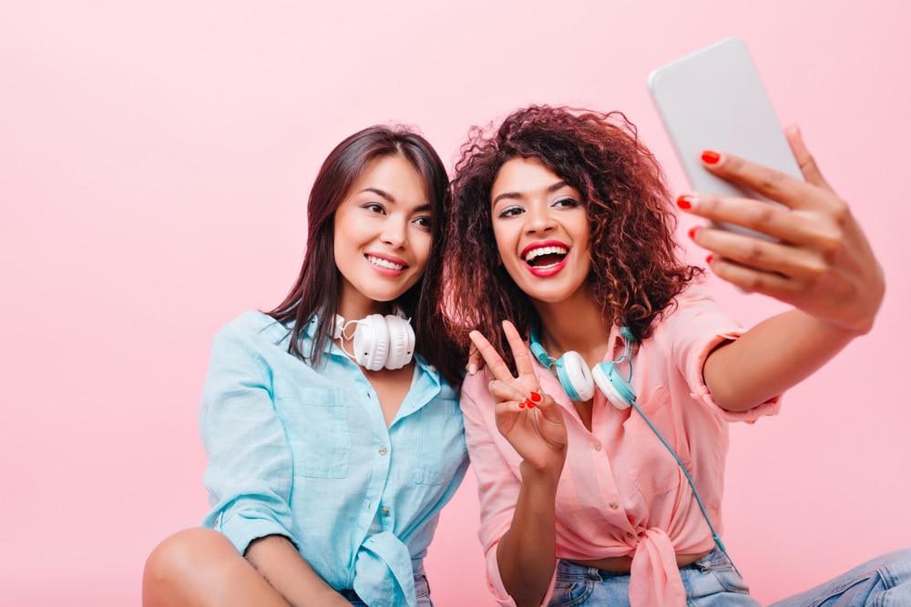 Two young women engaging in an early onset of selfie-taking on a pink background.